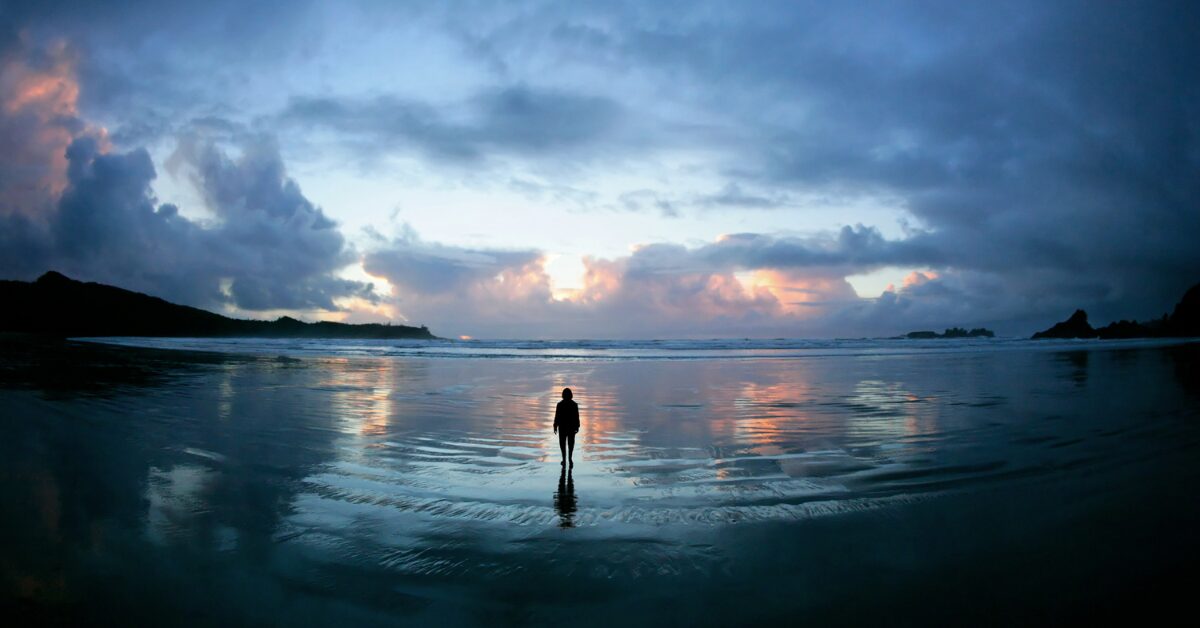Silhouette of a person standing alone on a vast reflective shoreline at sunrise, symbolizing clarity, space, and the journey to build a connected business ecosystem.